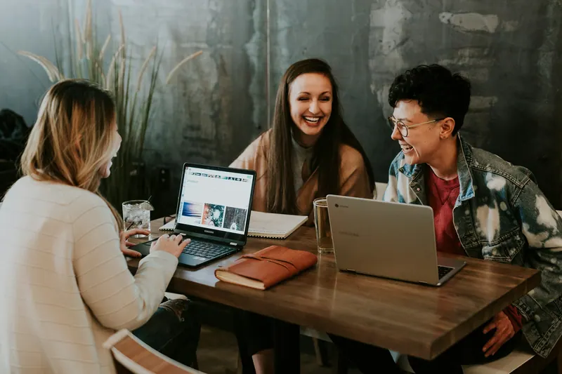 Three colleagues laughing around laptops at a wooden table, corporate lifestyle photography