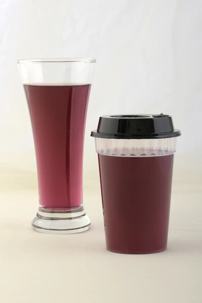 Dark berry juice in a tall glass beside a takeaway cup with a black lid on a soft grey studio backdrop, beverage photography in Pune
