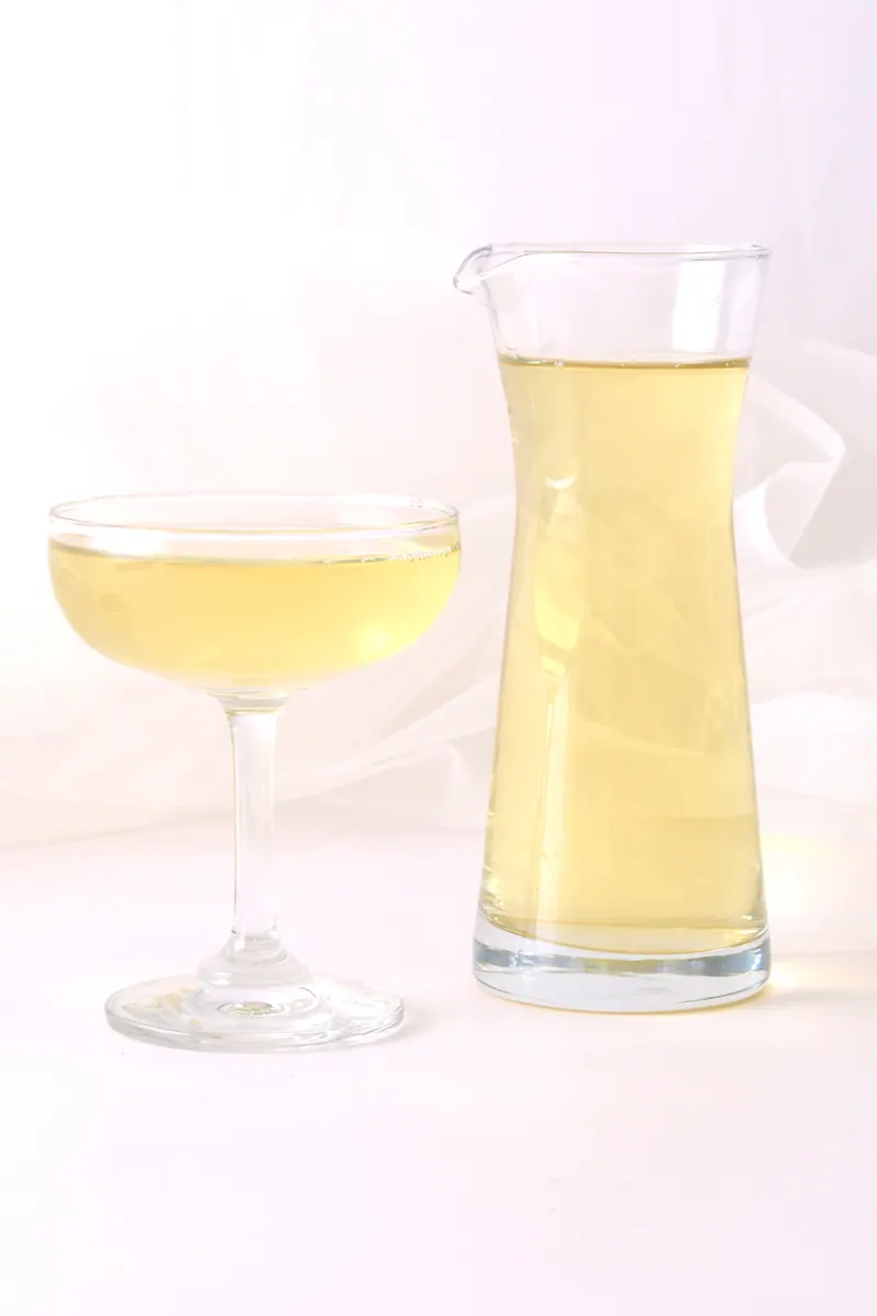 Pale golden drink in a stemmed glass beside a glass carafe against a soft lilac studio backdrop