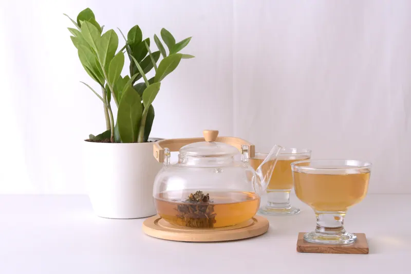 Glass teapot and cups of herbal tea beside a potted green plant on a white table, lifestyle food photography in Pune