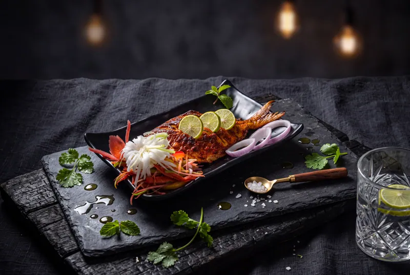 Grilled fish with onion rings and herbs on black slate beside a glass of lime water, restaurant food photography in Pune