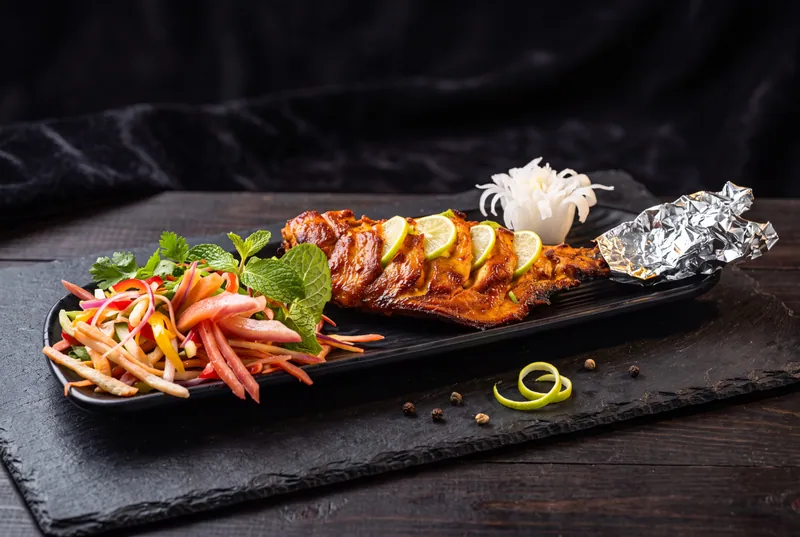 Grilled fish tikka with lime, salad and foil on a black slate board, dark moody restaurant food photography in Pune