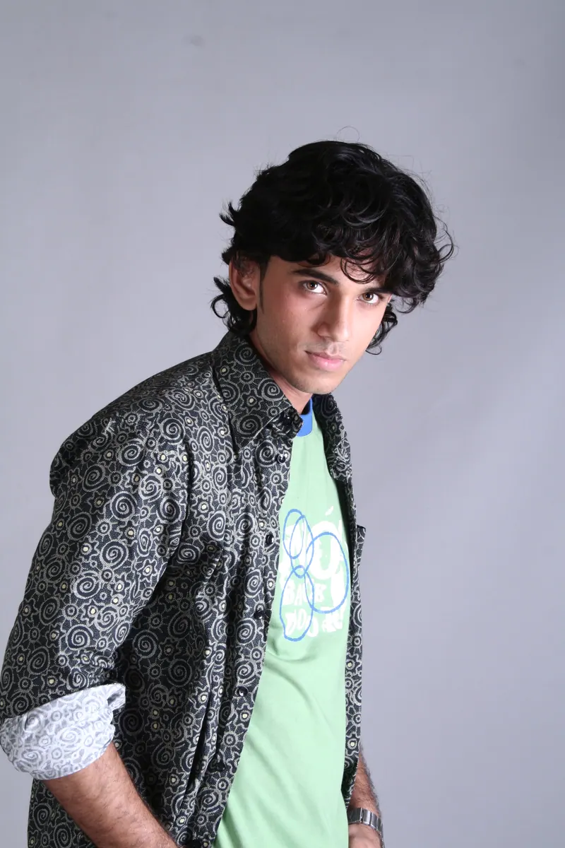 Young man with curly hair wearing a patterned jacket over a green graphic tee against a grey backdrop, editorial fashion photography
