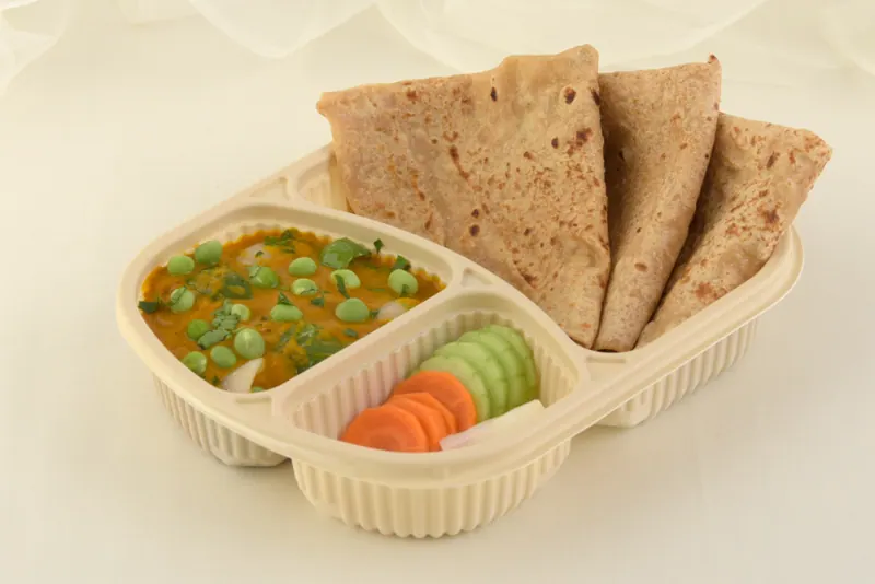 Takeaway meal tray holding green peas curry, folded chapatis and a cucumber carrot salad, food delivery packaging photography