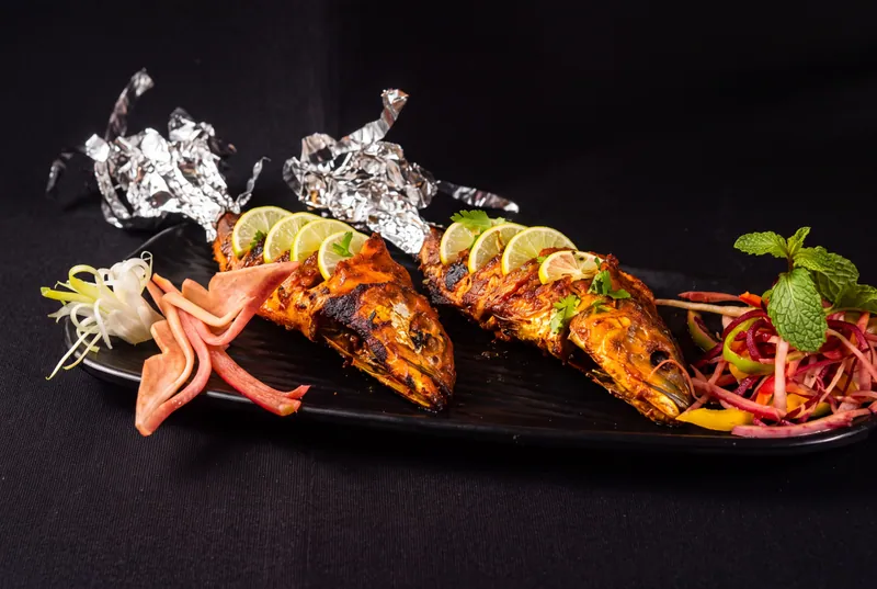 Tandoori fish with silver foil, lime wedges and a carved garnish on a dark backdrop, restaurant menu photography in Pune